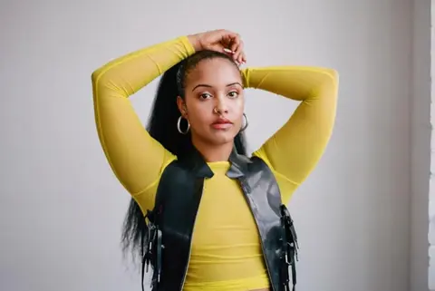 A person with long hair poses with arms raised, wearing a yellow long-sleeve top and a black vest, against a light background.