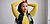 A person with long hair poses with arms raised, wearing a yellow long-sleeve top and a black vest, against a light background.