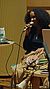 A woman with curly hair speaks into a microphone while seated at a table with electronic equipment and a decorative item.