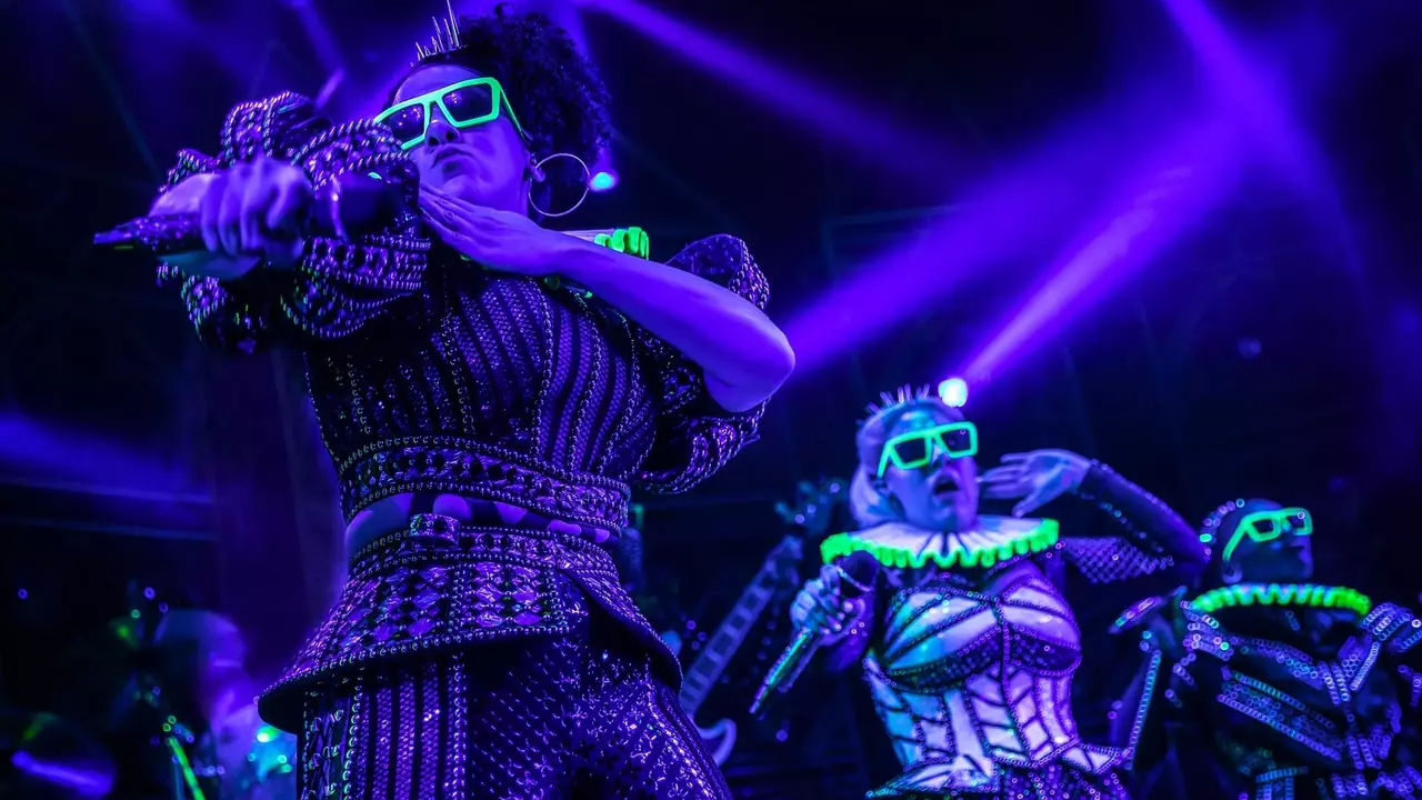 Three performers in vibrant, sequined outfits and neon accessories dance on stage under purple lighting, showcasing dynamic movements and synchronized choreography.
