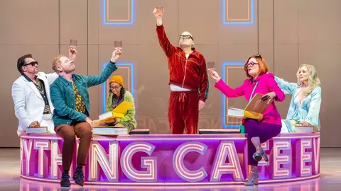 A group of six performers, dressed in colorful outfits, raise glasses in celebration around a circular stage with the text "Everything Can Be."
