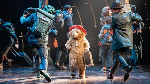 A bear in a red hat and suitcase stands center stage, surrounded by people with backpacks, in a theatrical setting.