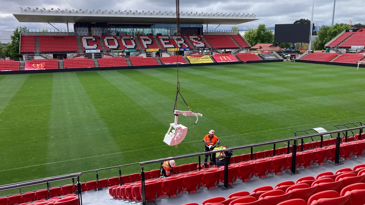 Workers in safety gear prepare seating at Coopers Stadium, with a well-maintained green pitch and empty red seats visible.
