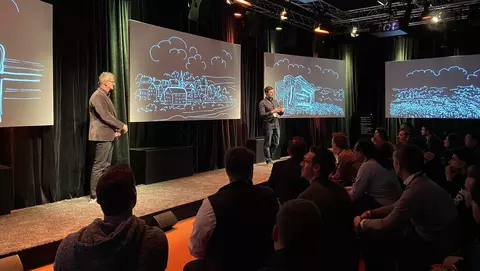 Two presenters stand on stage in front of large screens displaying illustrated cityscapes, addressing an audience seated in front of them.