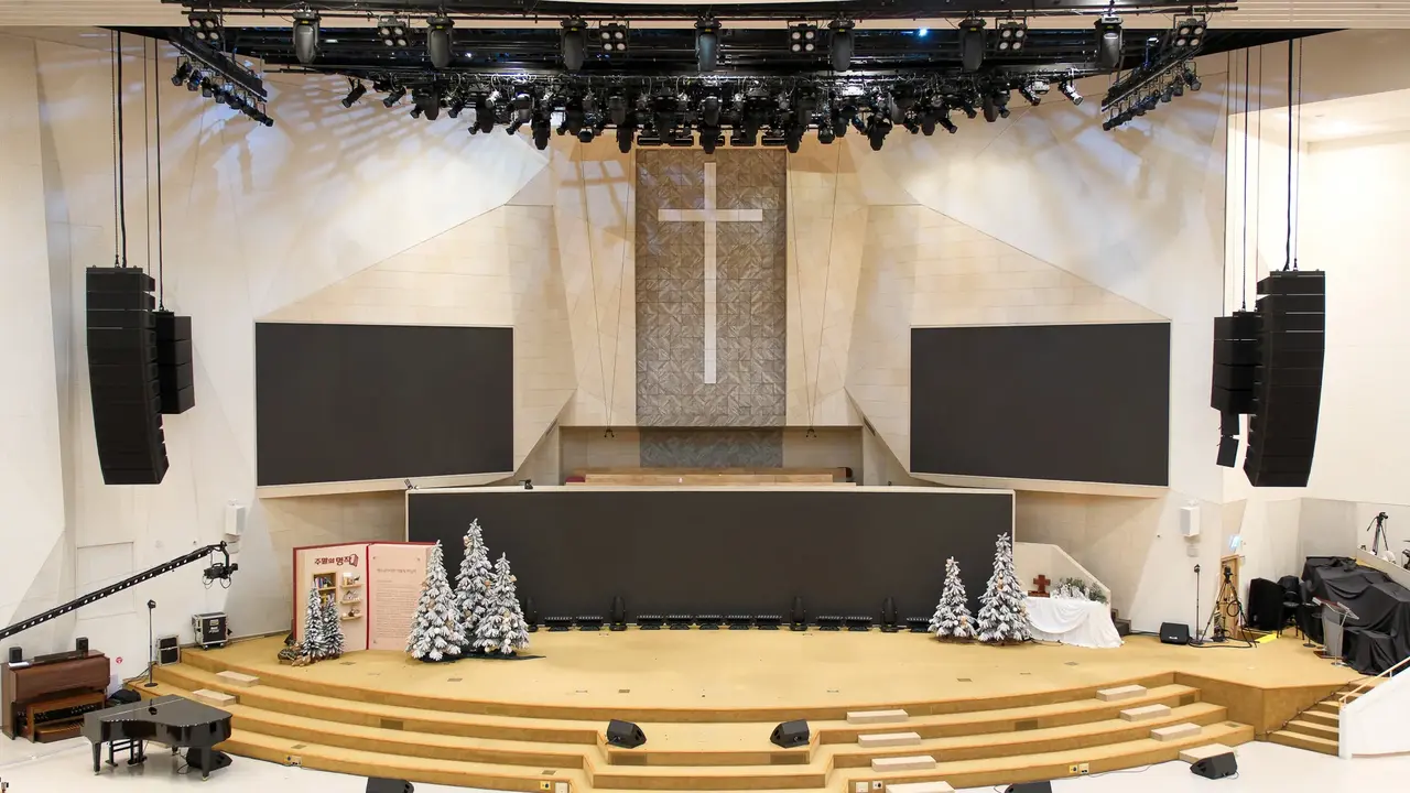 A modern church stage featuring a large cross, two blank screens, and Christmas decorations, with seating arranged in front.
