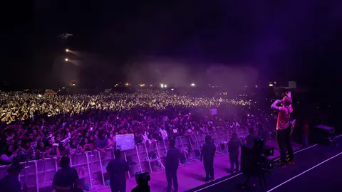 A performer stands on stage in front of a large crowd at a concert, with bright lights and a vibrant atmosphere.