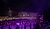 A performer stands on stage in front of a large crowd at a concert, with bright lights and a vibrant atmosphere.