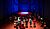 Audience gathered in a dimly lit performance space with a circular spotlight on the stage, surrounded by blue lighting.