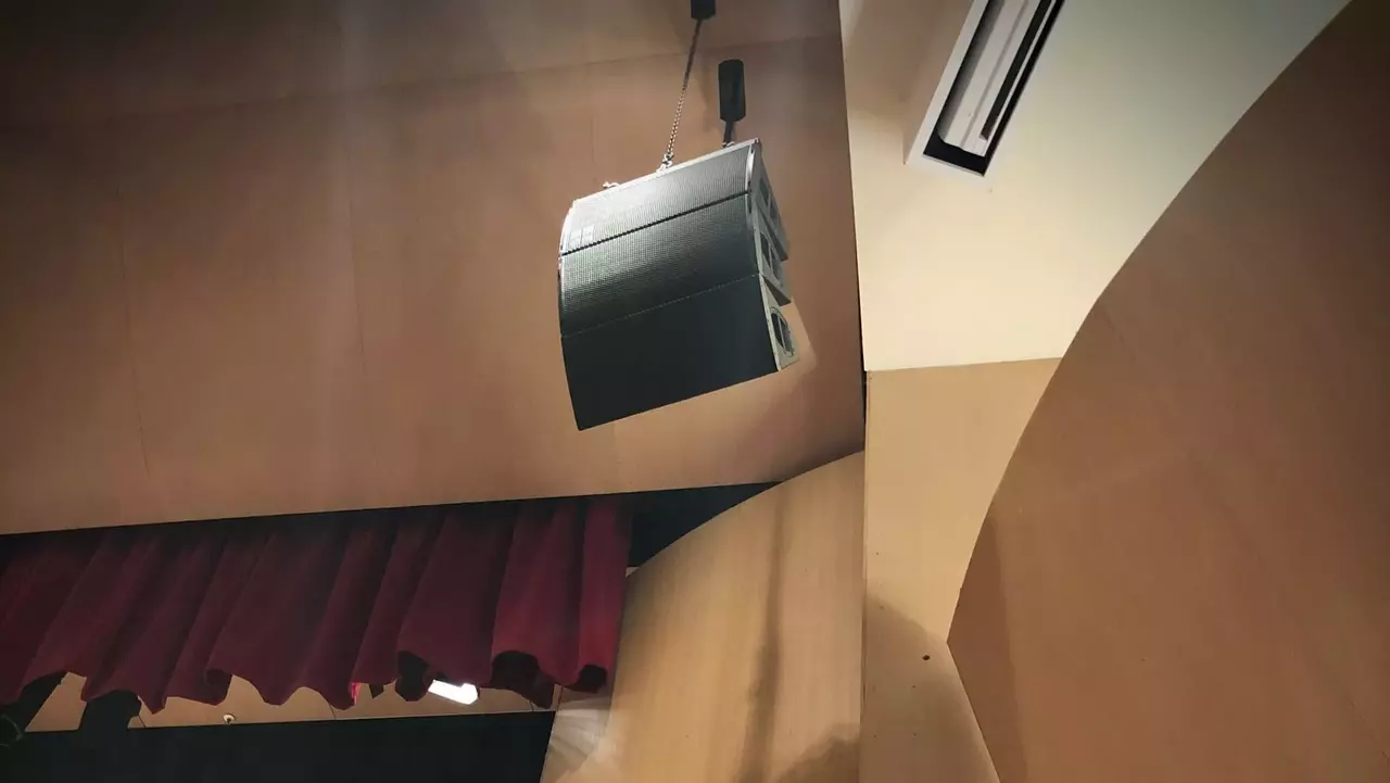 Suspended audio speaker in a modern auditorium, with red curtains and wooden paneling visible in the background.
