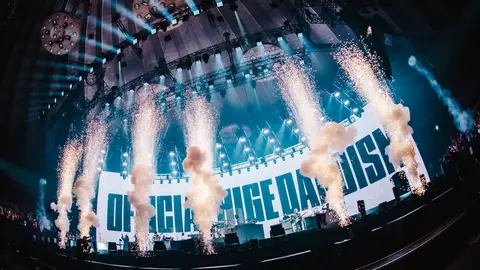 Concert stage with large backdrop displaying "OFFICIAL CHARGE DADIS" and pyrotechnics creating bursts of smoke and sparks.