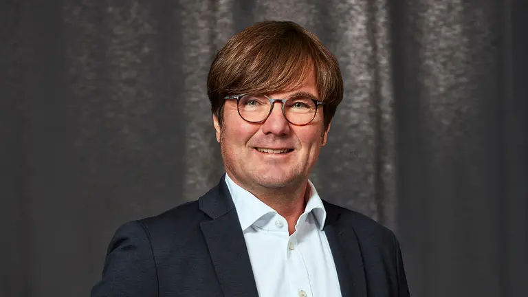 Man with glasses and brown hair, wearing a dark suit and white shirt, smiles against a gray backdrop.