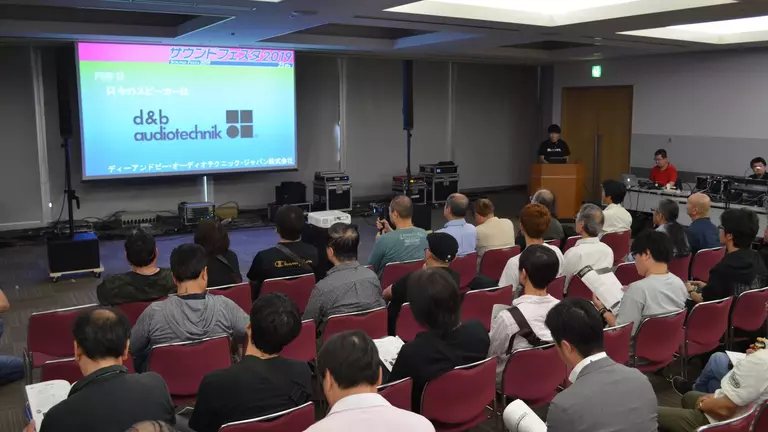 Audience seated in a conference room, facing a presentation screen displaying logos for d&b audiotechnik and other text.