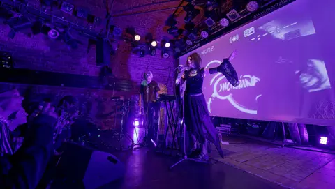 A performer in a flowing black outfit sings on stage, accompanied by a musician, with colorful lights and a projection screen in the background.