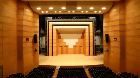 Empty auditorium with a wooden stage featuring a geometric backdrop and rows of black seats. The lighting is bright and focused on the stage area.