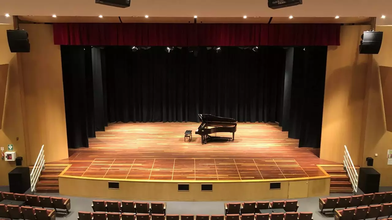 A stage with a grand piano set against a black curtain, surrounded by empty seating in a concert hall.