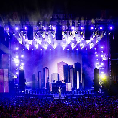 A performer stands on stage in front of a large crowd, illuminated by colorful lights and a backdrop of city skyline graphics.