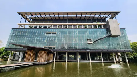 Modern glass building with a wooden entrance, elevated above a pond, featuring a cantilevered section and greenery on the roof.