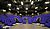 Theater interior featuring tiered seating with purple cushions, a central stage area, and overhead lighting fixtures.