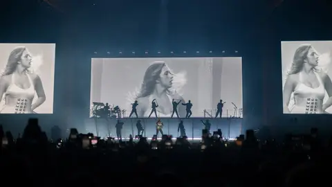 A performer on stage with a band, flanked by large screens displaying a black-and-white image of a woman, during a concert.