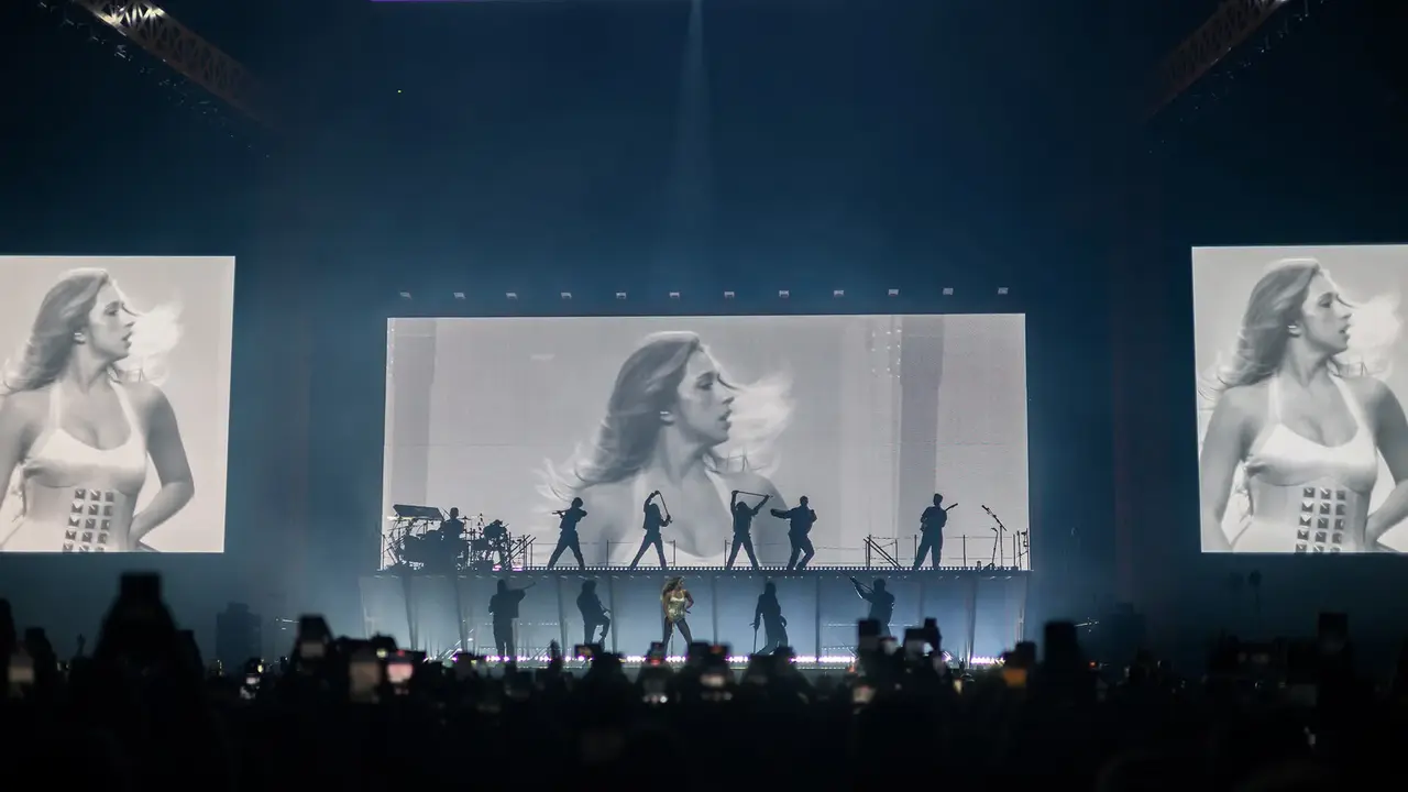 A performer on stage with a band, flanked by large screens displaying a black-and-white image of a woman, during a concert.