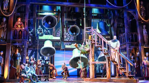 A theatrical scene featuring actors on a set with large bells and a staircase, depicting a vibrant performance with various characters.