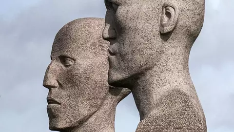 Two abstract stone sculptures of human heads, positioned closely together, overlooking a body of water under a cloudy sky.