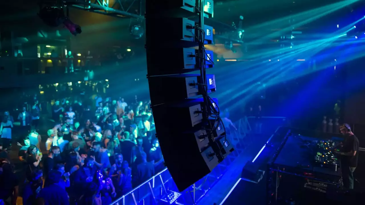 Ein DJ tritt auf der Bühne mit großen Lautsprechern und bunten Lichtern auf, während eine Menge die Musik in einem lebhaften Veranstaltungsort genießt.