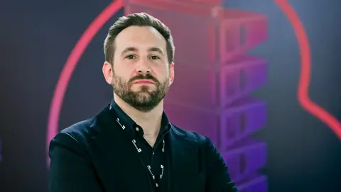 Man in a black blazer stands confidently with arms crossed, wearing a name badge, against a colorful abstract background.