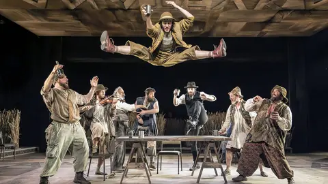 A performer leaps above a table surrounded by seven other actors, all dressed in period costumes, in a theatrical scene.
