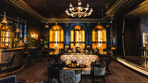 Luxurious bar interior featuring dark walls, gold accents, elegant chandeliers, and plush seating arrangements.