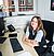 A young woman with long hair sits at a desk in an office, smiling. A computer keyboard, phone, and notepad are visible on the desk.