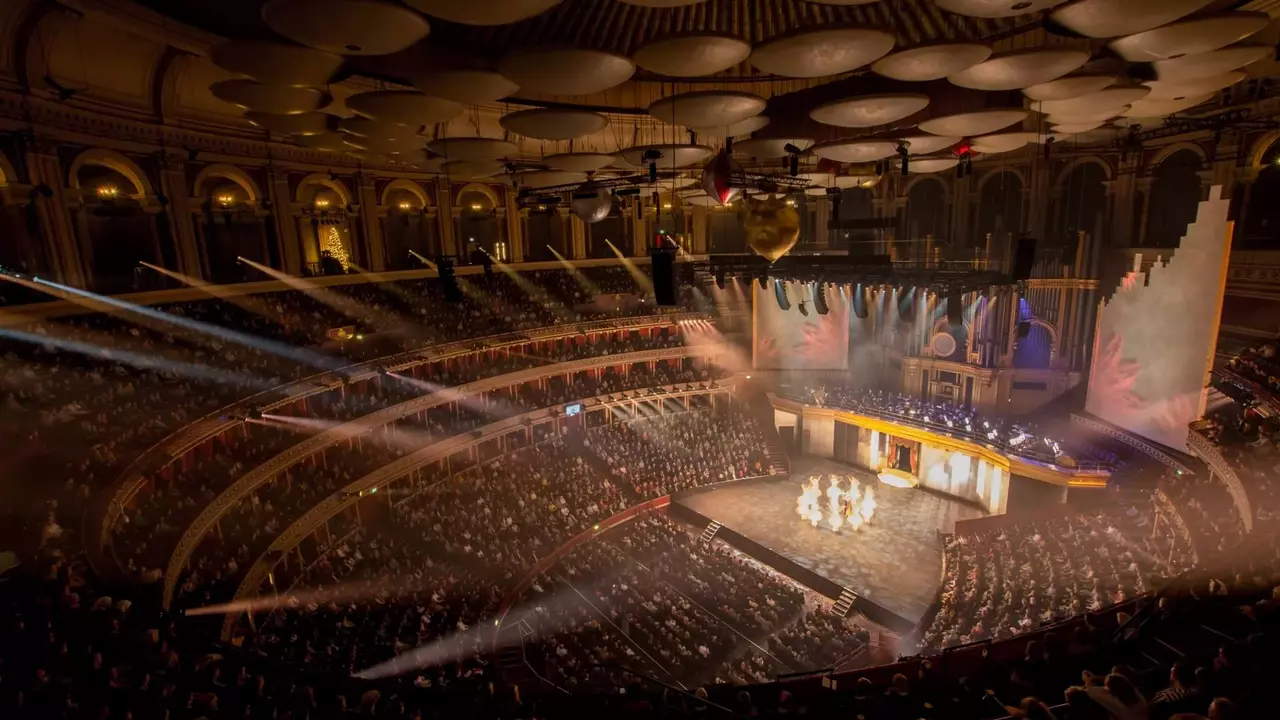 A large audience watches a performance on stage in a grand theater, illuminated by stage lights and surrounded by ornate architecture.