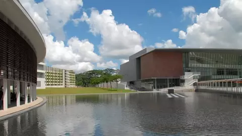 Modern architectural buildings reflect in a calm water surface, surrounded by green grass and a blue sky with fluffy clouds.