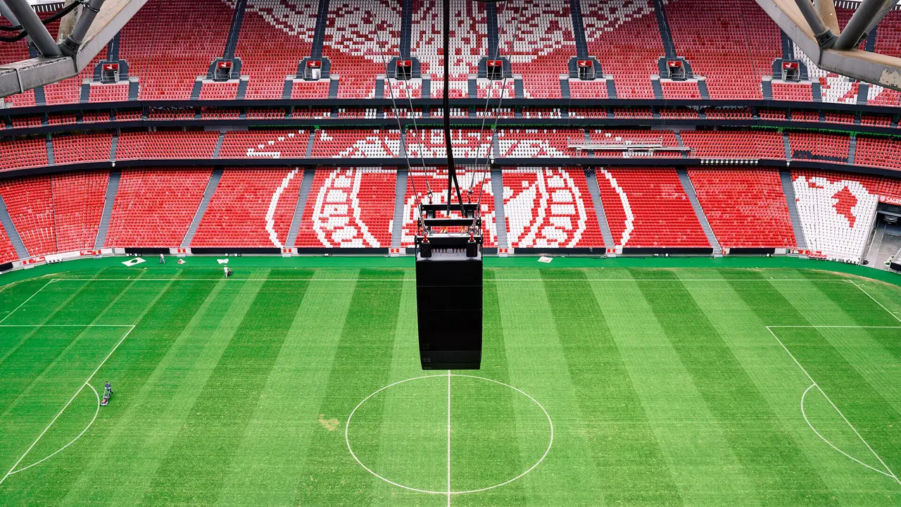 Aerial view of a soccer stadium featuring red seating and a central field, with a large scoreboard hanging above the pitch.
