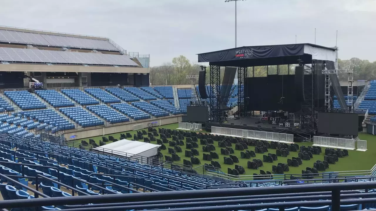 An outdoor stadium setup featuring a large stage, empty seating, and rows of chairs arranged on the field, under a cloudy sky.