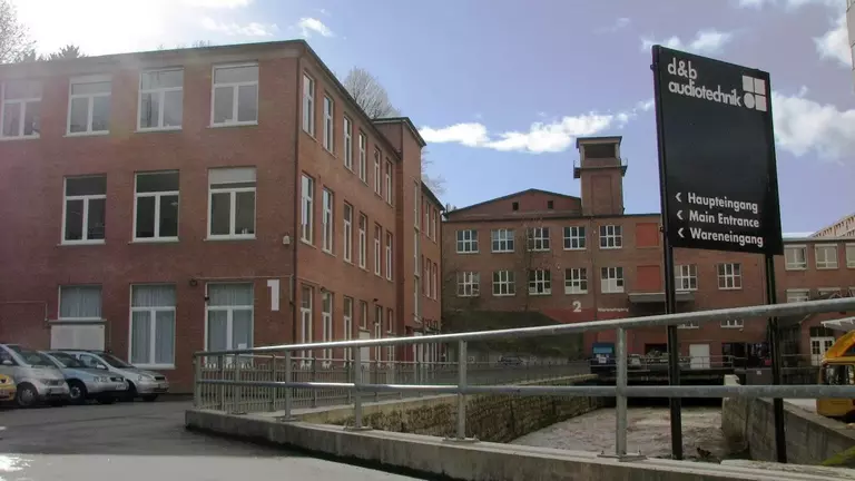 Red brick buildings with a sign for d&b audiotechnik, indicating the main entrance and visitor access. A parking area is visible in the foreground.