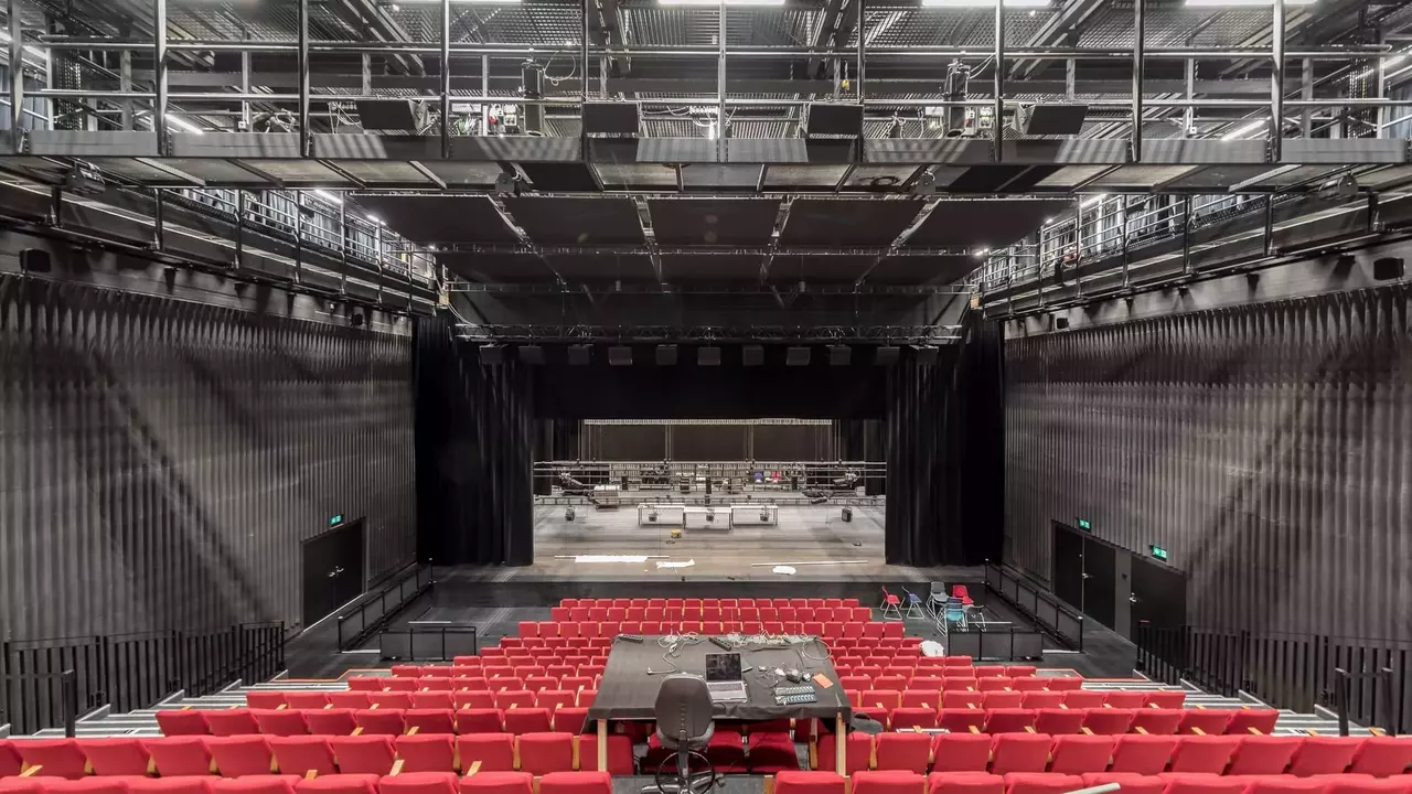 Red theater seats face a large stage with a black backdrop, surrounded by a modern, industrial-style auditorium.