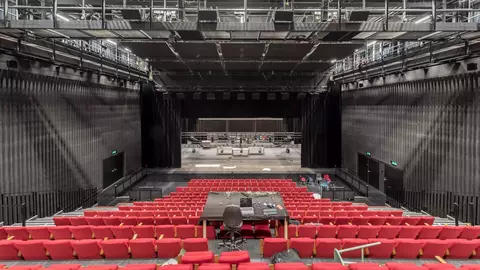Red theater seats face a large stage with a black backdrop, surrounded by a modern, industrial-style auditorium.