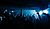 Crowd of people at a concert or club, with hands raised in the air, illuminated by blue and white lights.