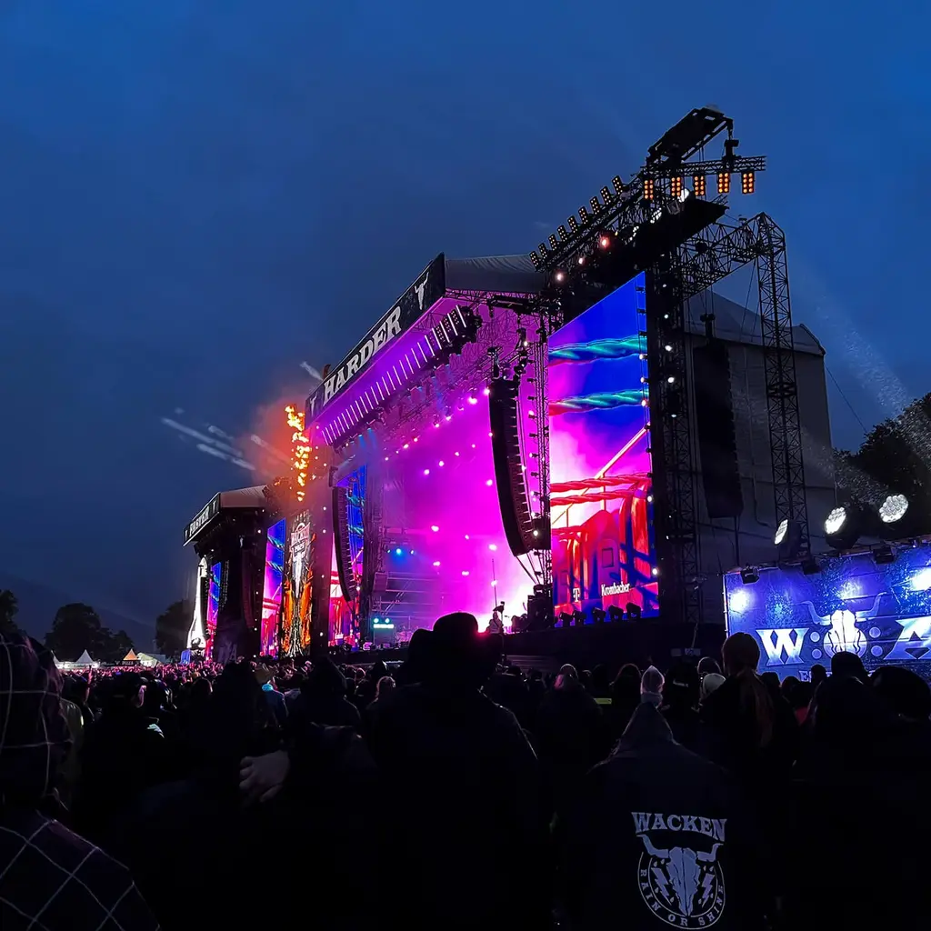 A large outdoor concert stage illuminated with colorful lights, surrounded by a crowd of attendees in dark clothing, under a twilight sky.