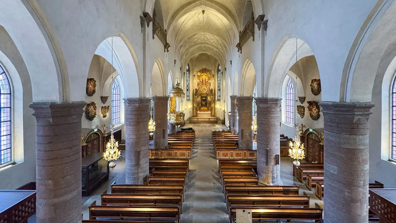 Interior view of a church featuring high arches, wooden pews, and a central altar, illuminated by chandeliers and natural light.