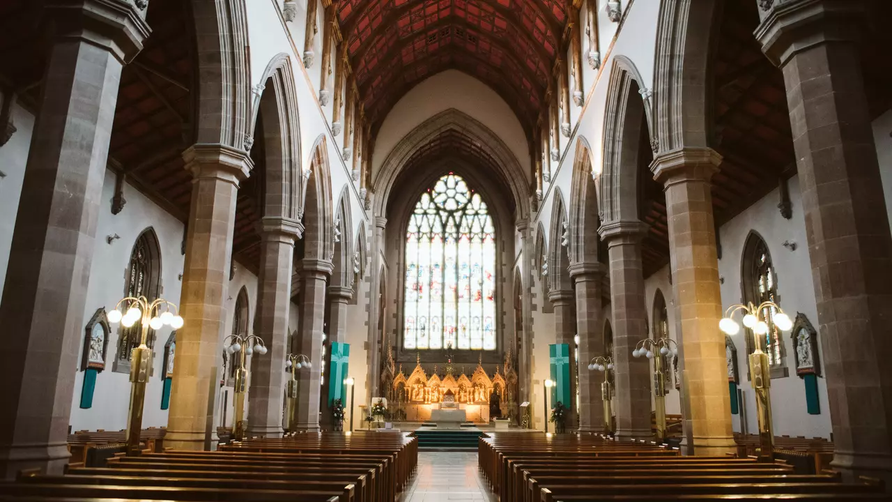 Interior of a church featuring wooden pews, tall arches, and a large stained glass window at the altar, illuminated by soft lighting.