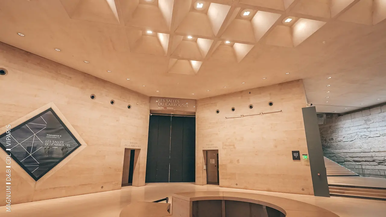 Modern architectural interior of a museum lobby featuring geometric ceiling design, stone walls, and two entrance doors.