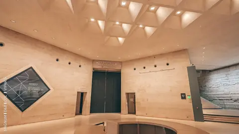 Modern architectural interior of a museum lobby featuring geometric ceiling design, stone walls, and two entrance doors.