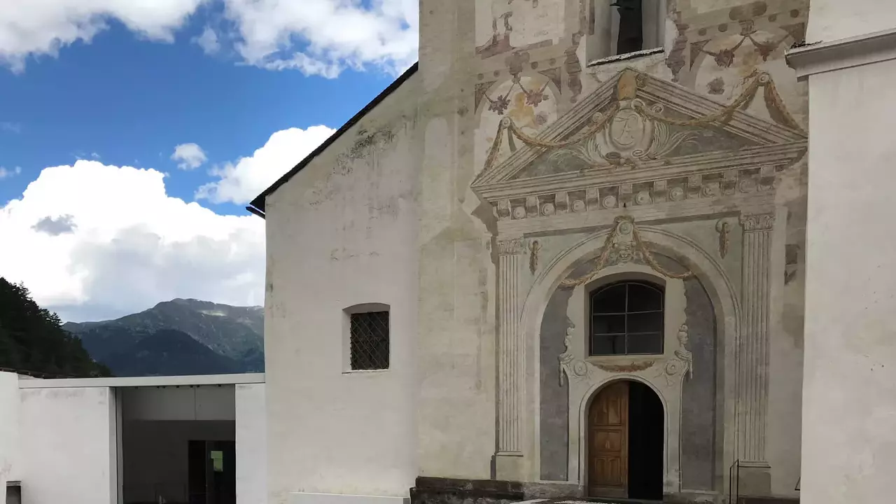 Historic church entrance featuring ornate frescoes and a statue, with modern architectural elements nearby against a mountainous backdrop.