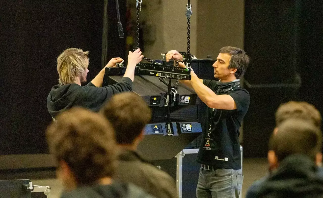 Two technicians are assembling equipment on a stage, with several people observing in the background.