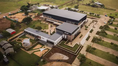 Aerial view of a modern building complex surrounded by landscaped grounds and open fields, featuring pathways and seating areas.