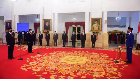 A formal ceremony with officials in suits standing in a decorated room, facing a speaker at a podium, with national flags displayed.