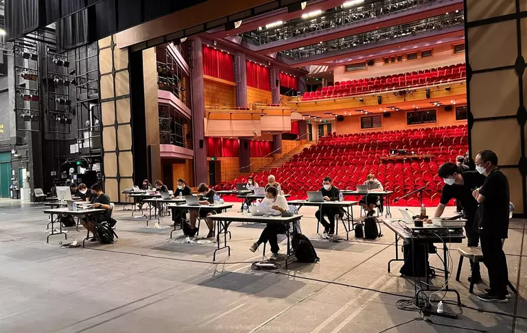 A group of people working at tables on a stage, with a red-seated auditorium visible in the background.