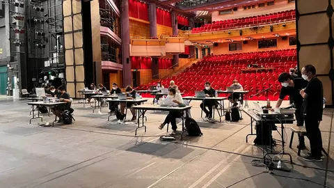 A group of people working at tables on a stage, with a red-seated auditorium visible in the background.
